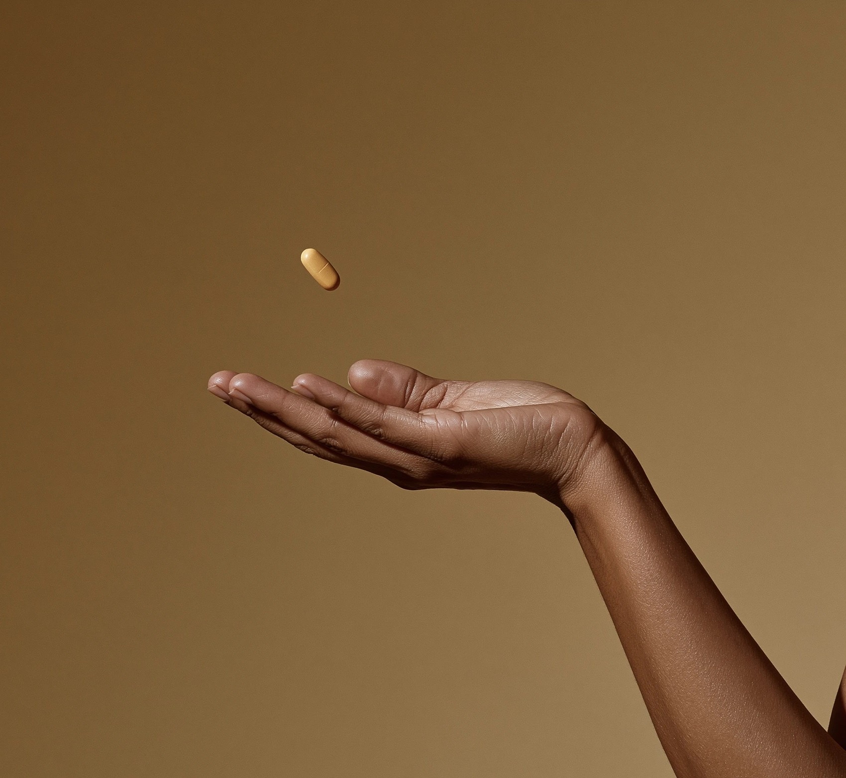 An open hand reaching up to catch a suspended GLP-1 capsule on a warm caramel backdrop